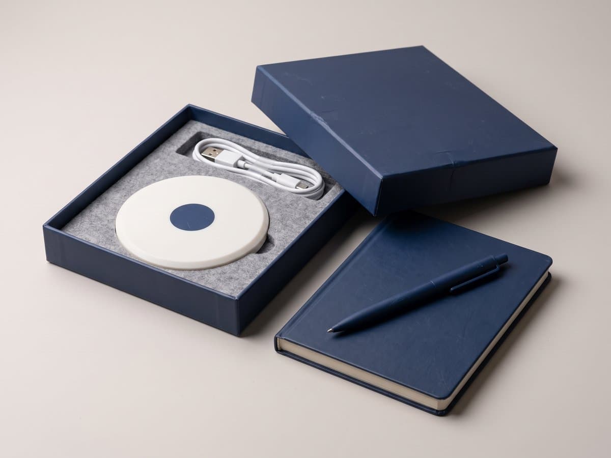 An open navy gift box holding a white charging pad marked with a navy circle, beside a navy notebook and pen.